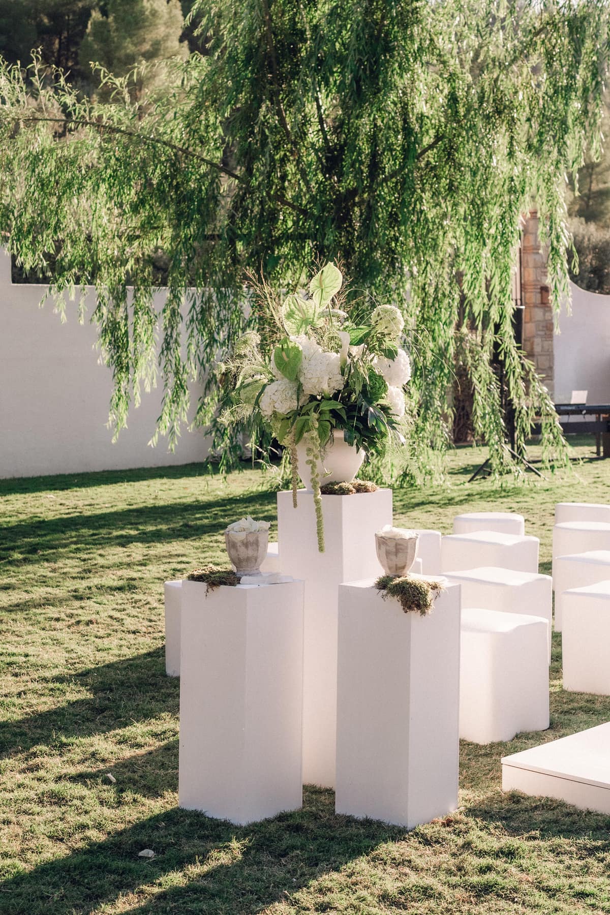 Decoración de ceremonia con pedestales blancos y flores bajo un árbol